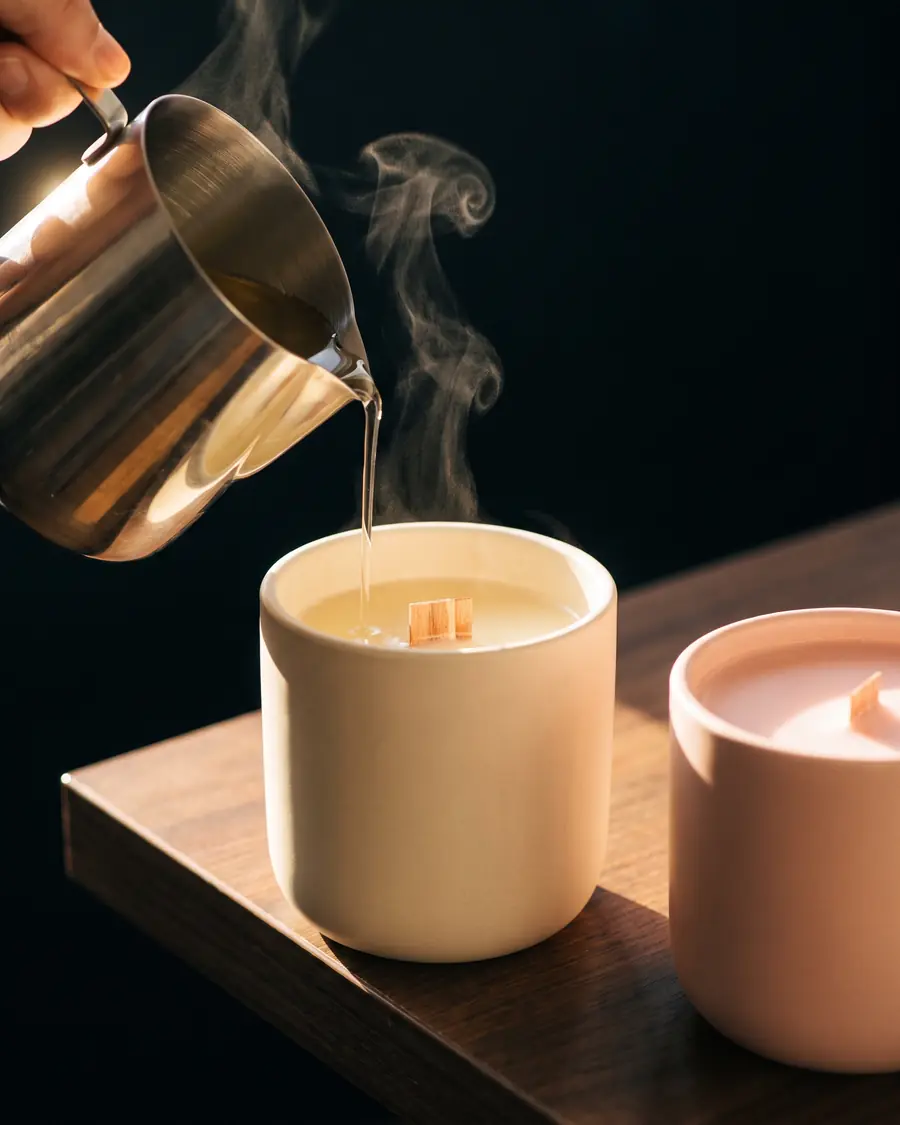 Molten soy wax being poured into a cream ceramic candle vessel beside a finished blush-pink candle
